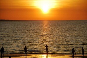 Ein Sonnenuntergang über einem Gewässer, im Vordergrund verschwommene Gestalten am Ufer.