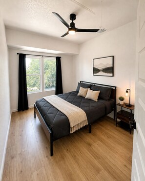 A bedroom with a bed, a ceiling fan, a window with curtains, a bedside table with a lamp, and a framed picture on the wall.
