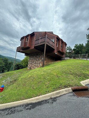 Una baita di legno con un balcone su una collina erbosa, circondata da alberi e un cielo nuvoloso.