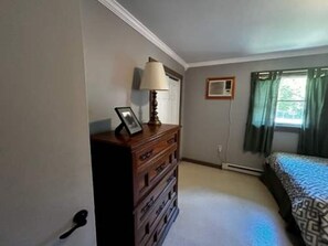A bedroom with a dresser, a lamp, a framed picture, a bed with patterned bedding, a window with curtains, and an air conditioning unit.