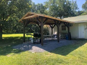 A covered outdoor area with a picnic table and grill, surrounded by a grassy lawn and trees.