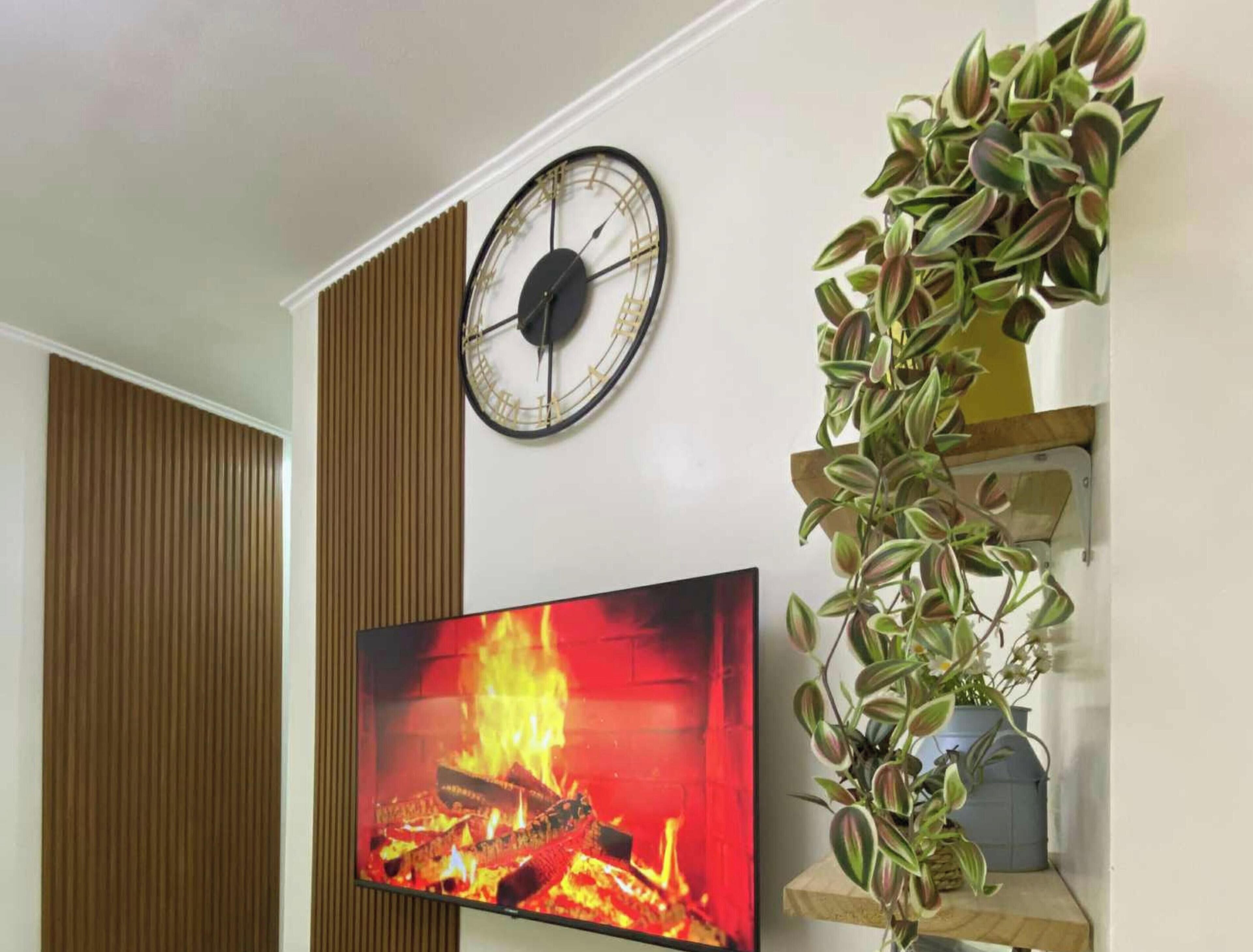 A room with a wall-mounted clock, a fireplace with a fire, and a plant on a shelf.