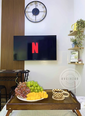 Ein an der Wand befestigter Fernseher zeigt das Netflix-Logo, eine Uhr und eine Ablage mit Früchten und einer karierten Tasse.