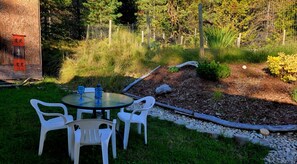 An outdoor seating area with a round table and white plastic chairs, surrounded by a garden with a wooden fence and greenery.