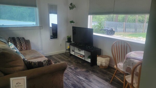 Sala de estar con sofá, televisor sobre un mueble bajo, una silla y ventana con vista a un coche y una cerca.