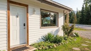A single-story house with a white exterior, a front door, a window, and a small garden area with rocks and plants.