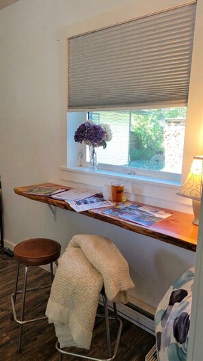 A small workspace with a wooden desk, a stool, a window with blinds, and a lamp.