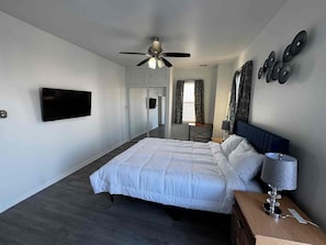 Ein Schlafzimmer mit einem Bett, einem Deckenventilator, einem an der Wand montierten Fernseher und einem Nachttisch mit Lampe.