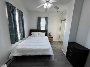 Une chambre à coucher comprenant un lit, une table de chevet, une commode, un ventilateur de plafond et une fenêtre avec des rideaux.