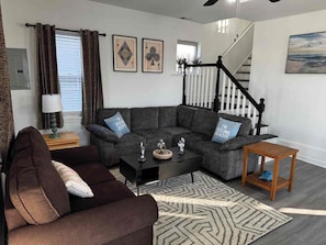 Ein Wohnzimmer mit einem Ecksofa, einem Couchtisch und einem Teppich. Eine Treppe mit Geländer und ein Gemälde an der Wand sind ebenfalls zu sehen.