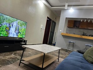 Ein Wohnzimmer mit Flachbildfernseher, einer Couch, einem Couchtisch und einer Küchenzeile mit Holdekor und Barhocker.