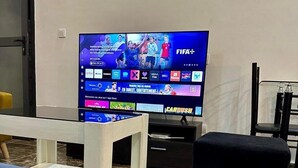 Ein Wohnzimmer mit einem Flachbildfernseher, der einen Sportkanal zeigt, einer Couch, einem kleinen Hocker und einem Glastisch.