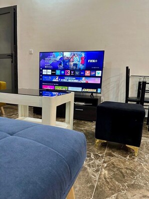 Ein Wohnzimmer mit einem Flachbildfernseher, der einen Sportkanal zeigt, einer Couch, einem kleinen Hocker und einem Glastisch.