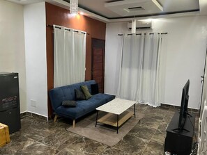 A living room with a blue sofa, a coffee table, a television, and a refrigerator. - Le Cocon de la Palmeraie - Quiet & Secure (Abidjan)