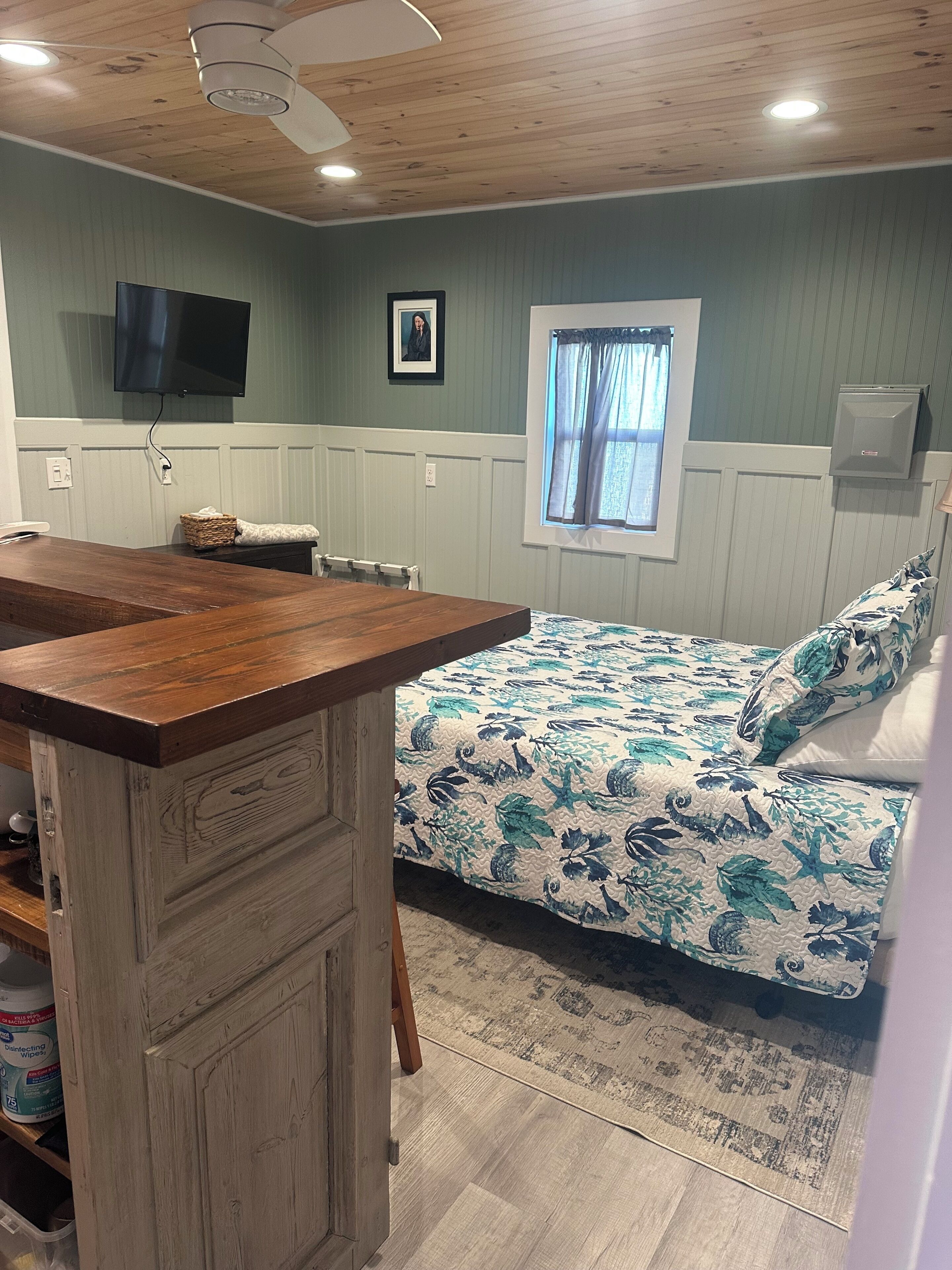 Une pièce avec un bureau en bois, un lit recouvert d’une literie à motifs floraux, un téléviseur fixé au mur et une fenêtre avec des rideaux.