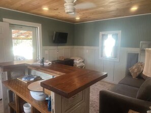 Une pièce avec un plafond en bois, un téléviseur fixé au mur, un canapé et un coin cuisine doté d’un plan de travail et d’un évier.