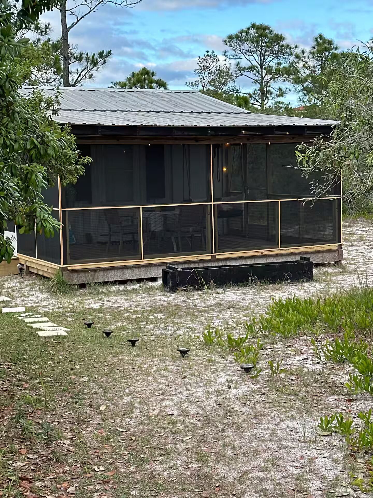 A small, single-story building with a metal roof, a screened porch, and a grassy area with scattered plants.