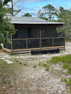 A small, single-story building with a metal roof, a screened porch, and a grassy area with scattered plants. - The weather will shine in October and November. Best beach time of the year. (Gulf Shores)