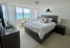 Un dormitorio con cama, mesitas de noche, escritorio y vista al mar.