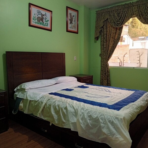 Un dormitorio con cabecera de madera, cama con colcha blanca y azul, dos cuadros en la pared y una ventana con cortinas.