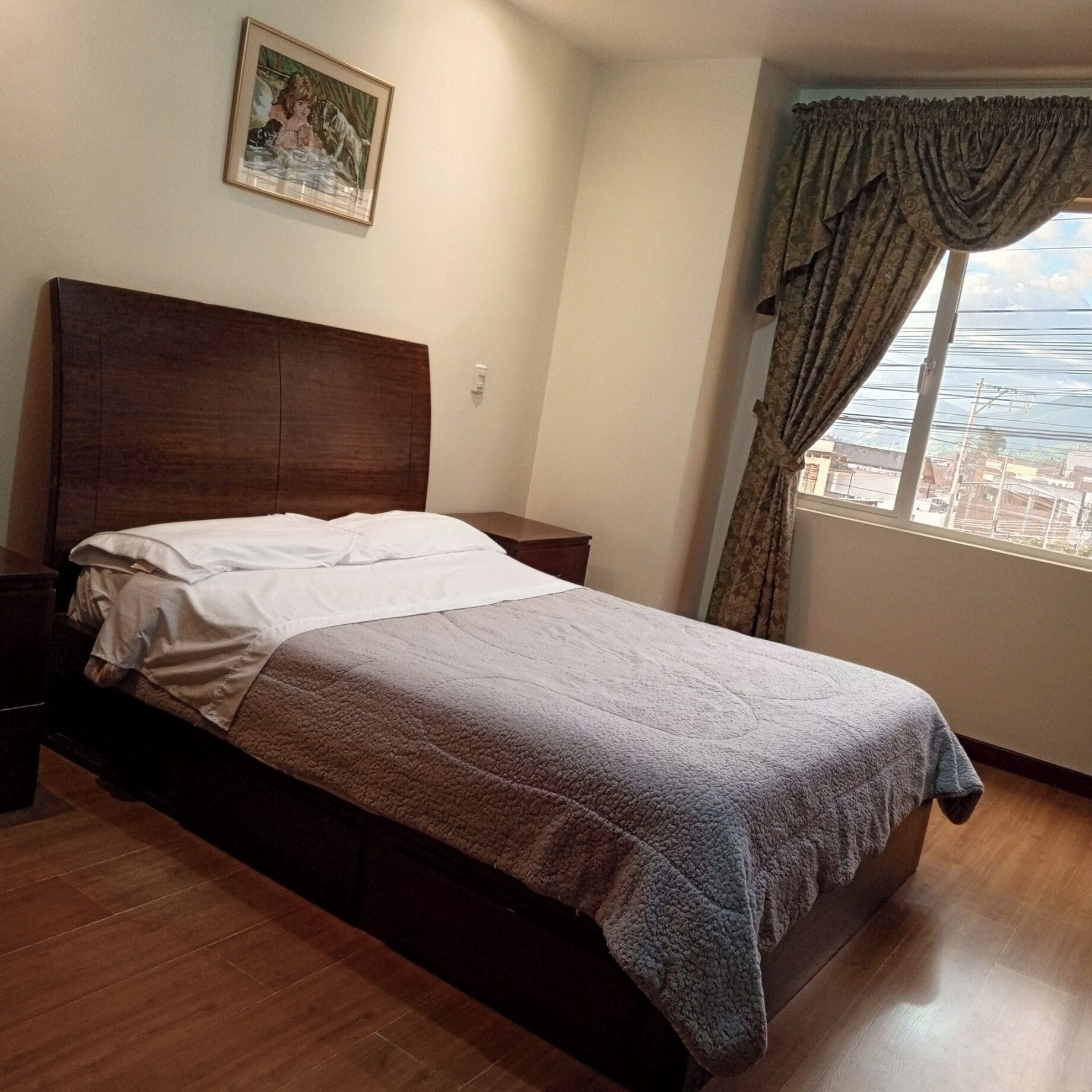 A bedroom with a wooden bed, a nightstand, a framed picture on the wall, and a window with curtains.