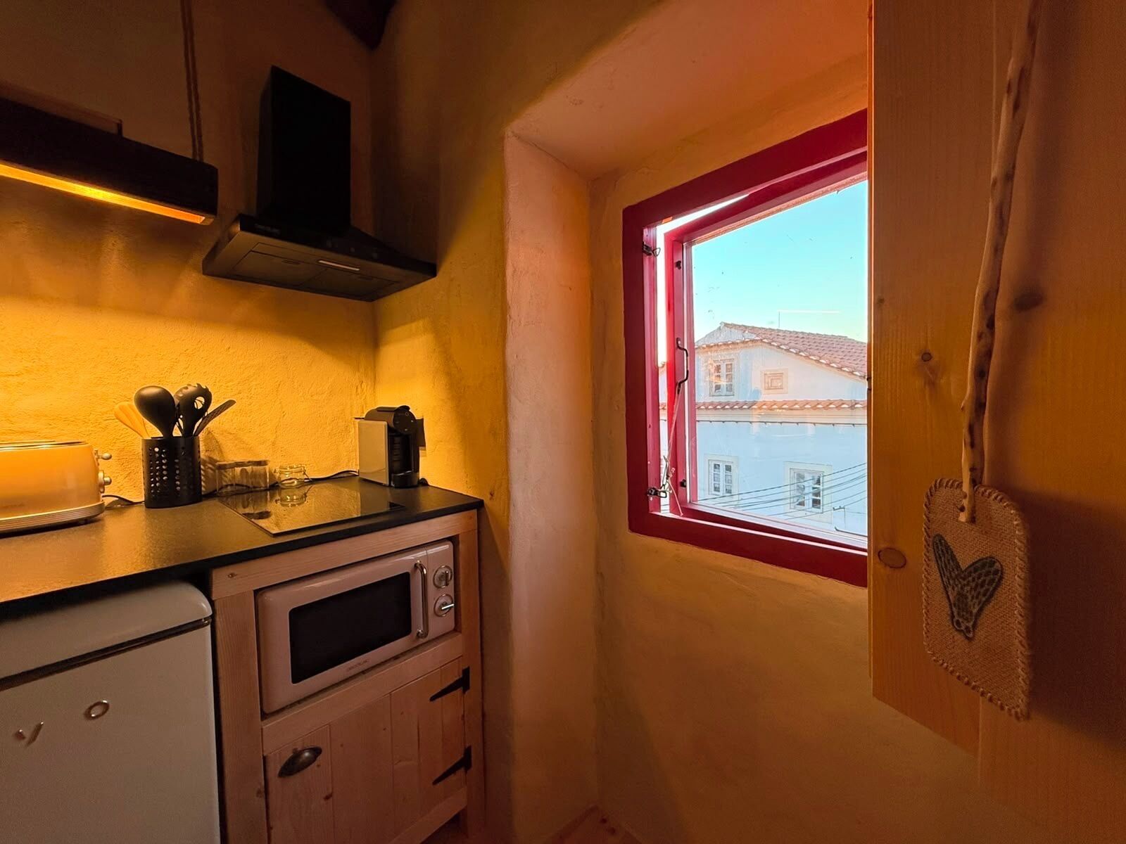 Cocina compacta con microondas, tostadora y cafetera sobre la encimera, una pequeña ventana con marco rojo y vistas a un edificio.