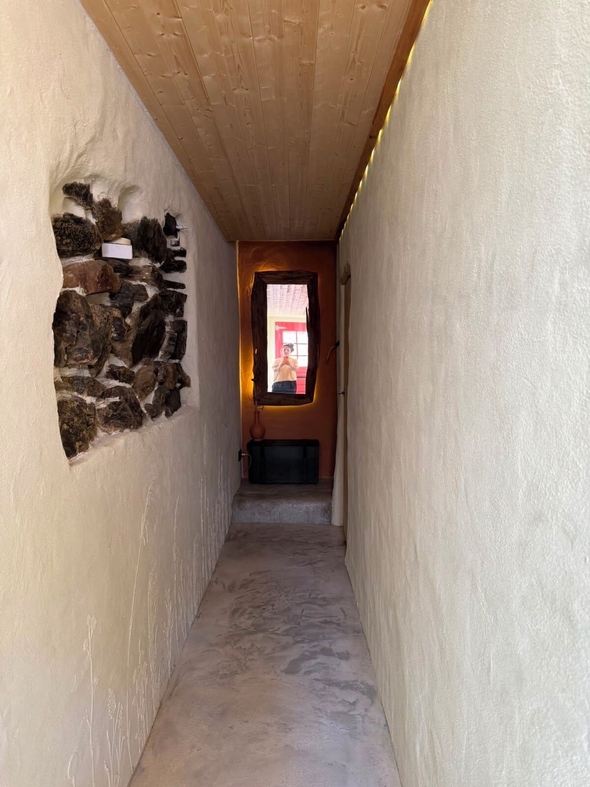 A narrow hallway with a stone wall on the left, a wooden ceiling, and a door at the end.