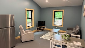A compact living space with a kitchenette, a flat-screen TV, a sofa, a dining table with chairs, and two windows with wooden frames.