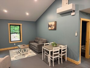 A room with a grey couch, a dining table with chairs, a window with a view of a forest, and an air conditioning unit.