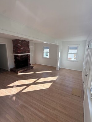 A spacious room with a brick fireplace, wooden flooring, and two windows. - Adorable 2-bedroom Seasonal Rental in Milford Beach Neighborhood (Milford)