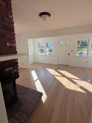 A spacious room with hardwood flooring, white walls, a brick fireplace, and multiple windows. - Adorable 2-bedroom Seasonal Rental in Milford Beach Neighborhood (Milford)