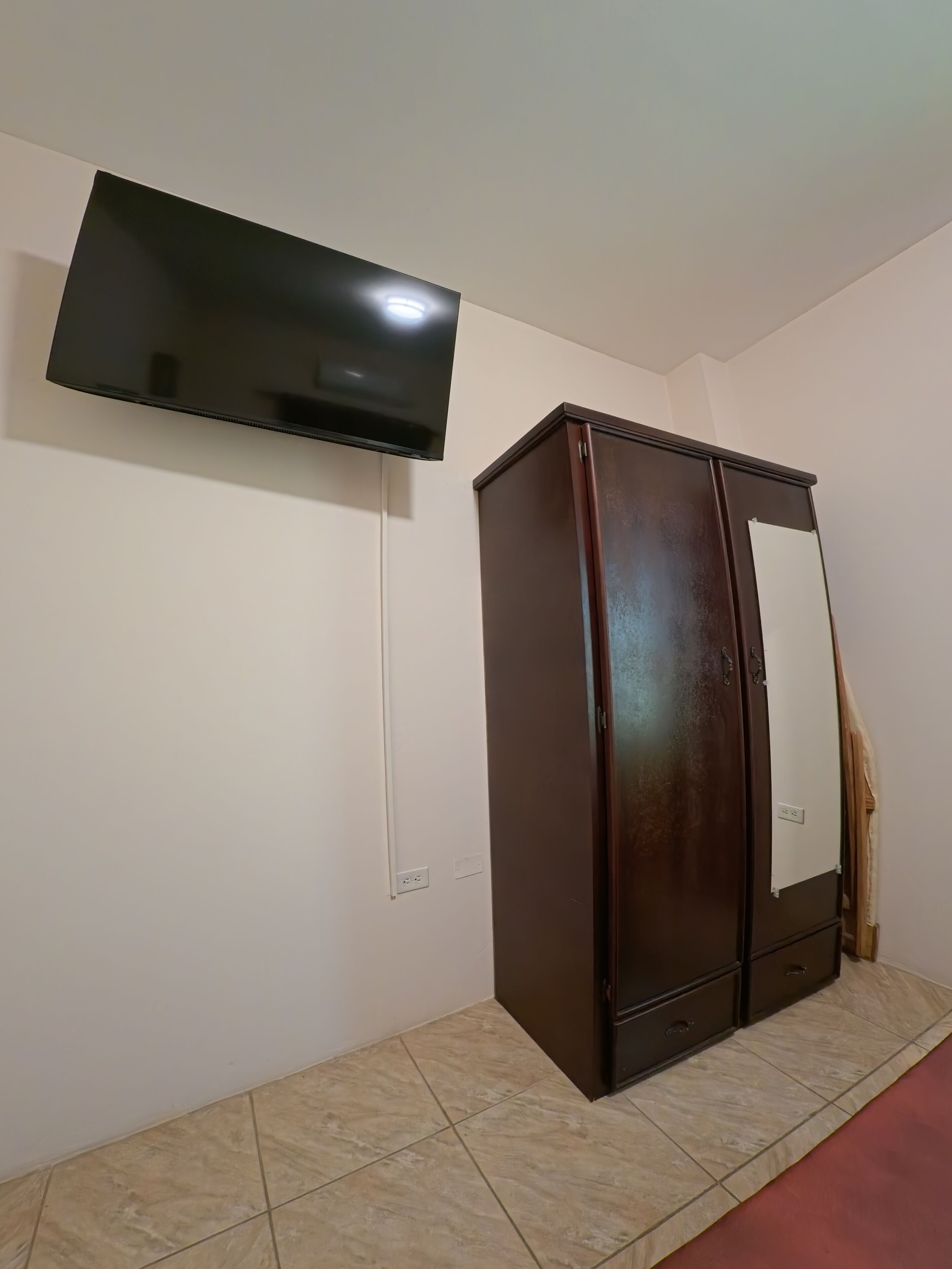 A room with a flat-screen TV mounted on the wall, a wooden wardrobe with a mirror, and a tiled floor.