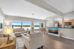Un vaste espace de vie avec une grande fenêtre donnant sur une plage, une cuisine équipée d’appareils électroménagers modernes et un coin salon confortable avec une lampe.