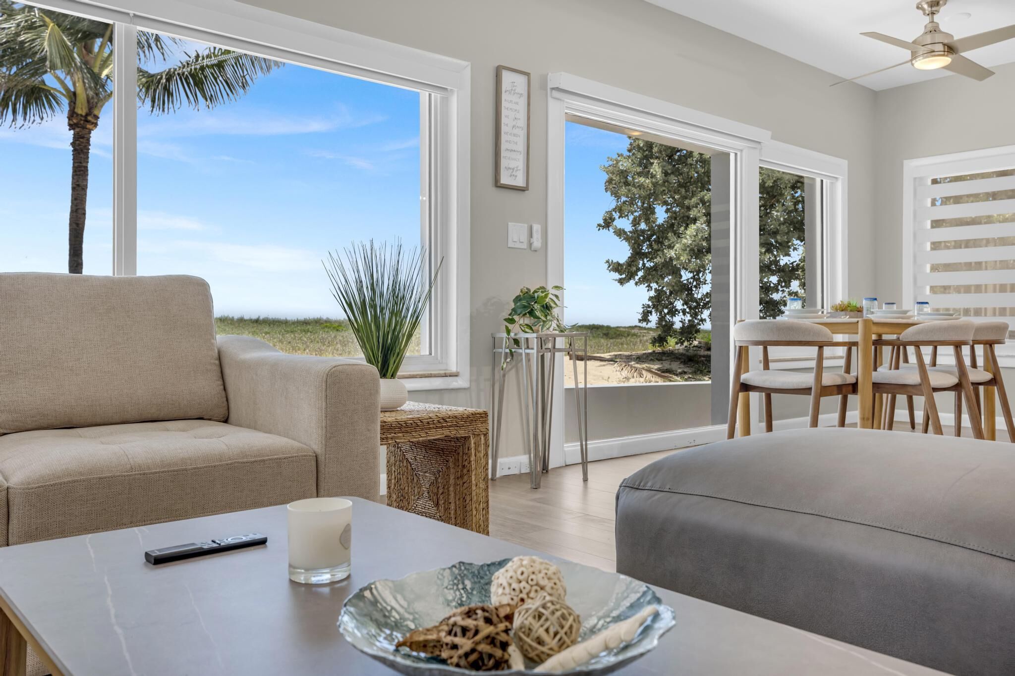 Een moderne woonkamer met een bank, een salontafel met decoratieve objecten, een eethoek met stoelen en grote ramen met uitzicht op het strand.