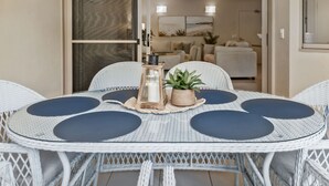 Uma mesa redonda com estrutura de vime branco e jogos americanos circulares escuros, acompanhada por uma pequena planta em vaso e um candeeiro.