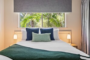 Une chambre d’hôtel avec un lit, deux lampes de chevet, une fenêtre donnant sur des palmiers et un rideau gris.