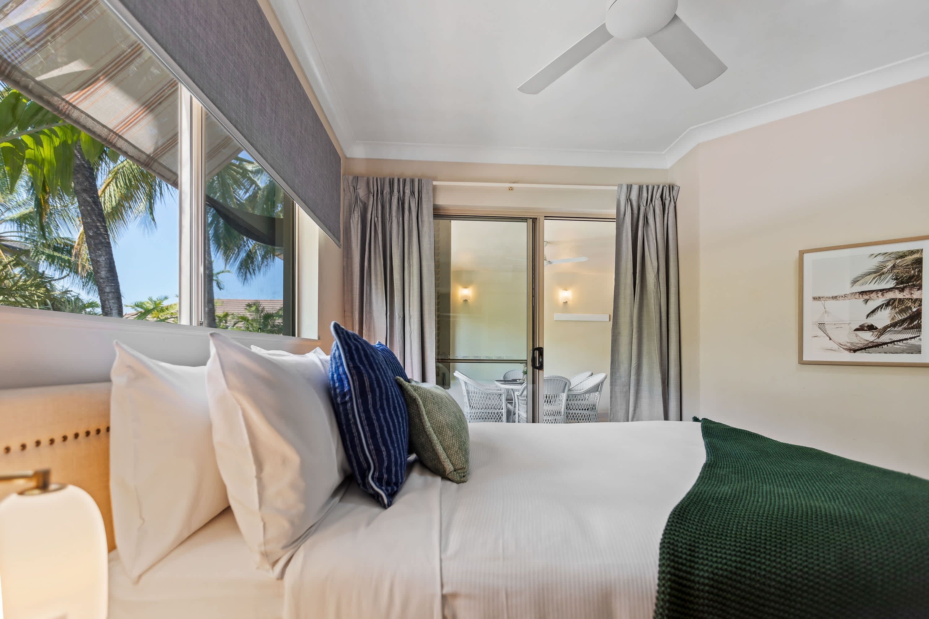 A hotel room with a bed, a ceiling fan, a view of palm trees, and a balcony with outdoor furniture.