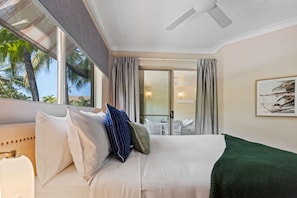 A hotel room with a bed, a ceiling fan, a view of palm trees, and a balcony with outdoor furniture. - Stunning Resort Apartment With Gym and Pool Access (Cairns)