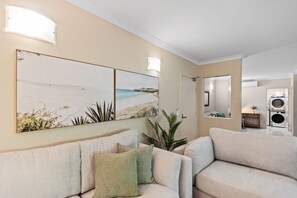 Un salon avec un canapé beige, un coussin vert et deux cadres représentant des scènes de plage, accrochés au mur.