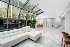 A modern living room with a sectional sofa, a dining area with a table and chairs, and a large glass window offering a view of palm trees. - Mega-Yacht Showcase Villa with Pool Table/Marble (Fort Lauderdale)