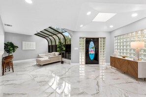 A modern interior with a marble floor, a sofa, a wooden sideboard, and a glass door with a tropical mural. - Mega-Yacht Showcase Villa with Pool Table/Marble (Fort Lauderdale)