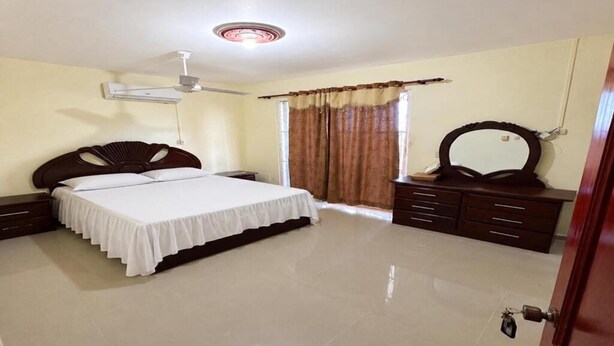 Quarto com cama, penteadeira, espelho, ventilador de teto e janela com cortinas.