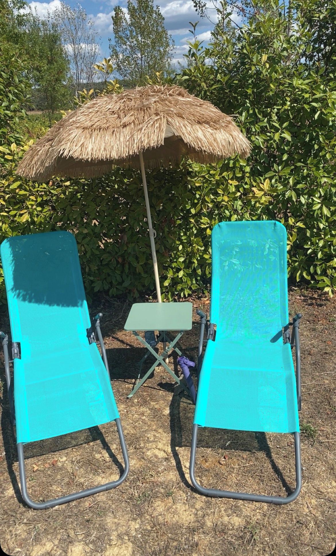 Two turquoise lounge chairs with a thatched umbrella and a small table in a garden setting.