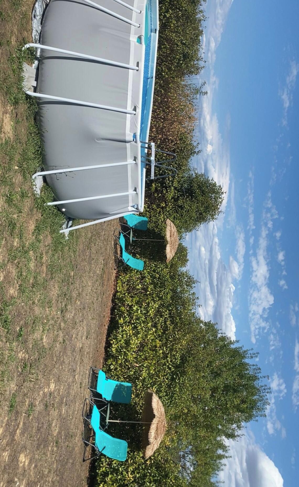 Una piscina con escalera, rodeada de césped y árboles. Hay dos sillas azules y un paraguas de paja.