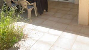 Un espace extérieur aménagé en terrasse, avec une table et des chaises, une porte-fenêtre coulissante donnant sur l’intérieur et un sol carrelé.