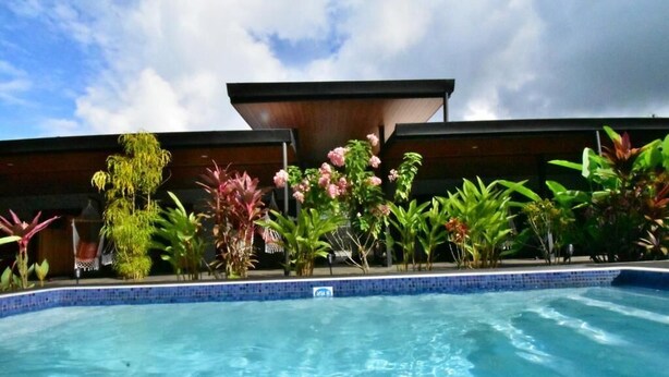 Une piscine dont le bord est recouvert de carreaux, entourée de diverses plantes tropicales, avec une structure de bâtiment moderne en arrière-plan.