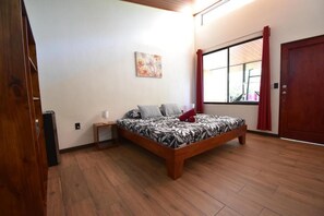 A bedroom with a wooden bed, a nightstand, a wall-mounted lamp, a window with curtains, and a door. - Raya (Matapalo)