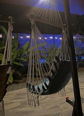 A hammock chair with a netted base, set against a backdrop of a pool and plants.
