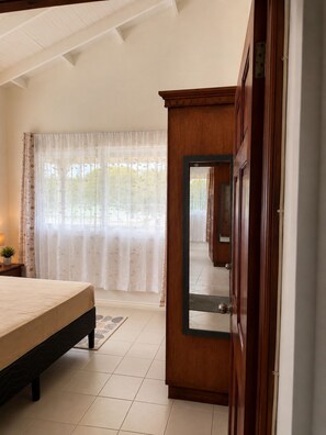 A bedroom with a bed, a wooden wardrobe, a window with sheer curtains, and a view of a garden. - Maison Teranga- 70m2, 2-BRM apartment, with AC, 5 minutes drive to Rodney Bay (Gros Islet)