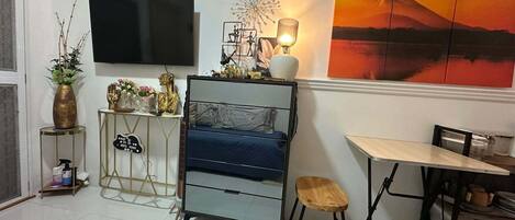 Una sala moderna con un televisor de pantalla plana, una cama con colchón azul, una mesa de comedor con sillas y un cuadro grande de un paisaje montañoso.